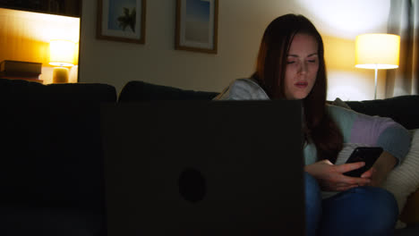 Mujer-Sentada-En-Un-Sofá-En-Casa-Por-La-Noche-Transmitiendo-O-Viendo-Películas-O-Programas-En-Una-Computadora-Portátil-Y-Navegando-Por-Internet-En-El-Teléfono-7